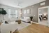 Bright and inviting living room with modern decor, featuring a cozy white sofa, sleek armchair, and a stylish ottoman table on a textured rug. Abundant natural light fills the room through large windows, complemented by elegant furnishings and serene wall art.