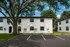 Charming apartment building with a classic white facade, nestled under a beautiful large tree, offering ample parking space in a serene neighborhood.
