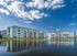 Charming lakeside apartment complex with modern blue and white facades, reflected in tranquil waters beneath a bright, cloud-dappled sky.