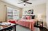 Bright and inviting bedroom featuring a cozy pink quilted bed, elegant black and white floral artwork, and a well-lit study area by a large window.