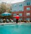 Charming apartment complex with vibrant red brick exterior, featuring a refreshing pool area complete with lounge chairs and colorful umbrella, exuding a lively and welcoming atmosphere.