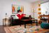 Vibrant living room featuring a plush black sofa, colorful abstract art, and a cheerful mascot in a red coat and top hat, adding a whimsical touch. Bright yellow and white curtains accentuate natural light, and a patterned rug completes the cozy ambiance.