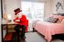 Charming bedroom with a light-filled window, cozy workspace with a quirky figure in a red outfit, and a stylishly adorned pink bed.