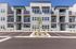 Modern apartment complex with sleek gray facade, multiple balconies, and ample parking space, surrounded by a landscaped garden.