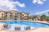 Charming apartment complex featuring a sparkling outdoor pool surrounded by modern lounge chairs and stylish patio seating, under a bright blue sky.