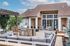 Charming outdoor patio with elegant seating, built-in stone grill, and lush greenery under a clear blue sky.
