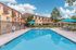 Inviting outdoor pool area with crystal-clear water, surrounded by stylish sun loungers and vibrant blue umbrellas, set against a charming residential building backdrop under a bright blue sky.