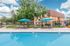 Inviting apartment complex with a sparkling pool, stylish loungers, and vibrant blue umbrellas set against a lush green backdrop.