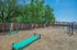 Outdoor fitness area with vibrant green exercise equipment, surrounded by lush trees and a wooden fence under a clear blue sky.