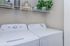 Modern laundry room featuring a sleek white washer and dryer set under a shelf adorned with plants and storage containers.