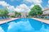 Inviting swimming pool area with sun loungers under a clear blue sky, surrounded by lush greenery and charming residential buildings.