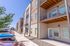 Modern apartment complex with private balconies, shaded in bright sunlight, offering ample parking and lush surroundings.