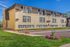 Charming three-story apartment building with beige exterior and modern shingles, surrounded by lush green grass and vibrant flower beds under a clear blue sky.