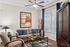 Cozy living room with elegant decor, featuring a plush sofa with blue cushions, stylish armchair, artistic wall painting, and a modern ceiling fan.