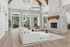 Elegant living room with a high ceiling, featuring a rustic stone fireplace, large windows overlooking greenery, a plush white sofa, and a grand chandelier.