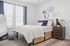 Bright and cozy bedroom featuring a comfortable bed with plush cushions, elegant nightstand with chic decor, and large windows allowing natural light to enhance the welcoming ambiance.