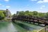Scenic view of a charming bridge over a serene green river, surrounded by lush trees and stylish apartment buildings under a bright blue sky.