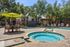 Inviting outdoor pool area with a bubbling hot tub, surrounded by lush trees and modern apartment buildings, featuring a shaded seating area with a vibrant green umbrella.