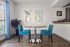 Charming dining area with stylish blue chairs and round wooden table, enhanced by abstract wall art and natural light through large window.
