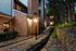 Charming evening view of a serene apartment pathway with elegant brick exteriors, illuminated by warm lighting and surrounded by lush greenery and stone landscaping.