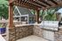 Charming outdoor patio with rustic wood pergola and modern stone barbecue grill, surrounded by lush greenery.