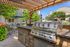 Charming outdoor patio with a sleek stainless steel grill and shaded seating area under a pergola, surrounded by lush greenery.
