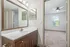 Chic bathroom with modern wooden vanity and large mirror, adjacent to a bright bedroom featuring plush carpeting and a picturesque window view.