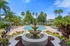 Luxurious apartment complex pool area featuring a grand fountain, surrounded by lush palm trees and elegant lounge chairs under a clear blue sky.