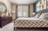 Elegant bedroom featuring a king-sized bed with patterned duvet, chic wooden dresser with decorative mirror, and large windows allowing natural light to flood the room.