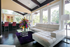 Bright, airy living space featuring a luxurious white sectional, vibrant floral centerpiece on a glass coffee table, and elegant floor-to-ceiling windows overlooking lush greenery.