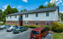Charming two-story apartment building with a landscaped front and ample parking, nestled amidst lush greenery under a clear blue sky.