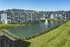 Modern apartment complex with elegant architecture, overlooking a serene pond with a fountain, surrounded by lush greenery.