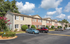 Charming row of townhouses with brick and siding exteriors, surrounded by lush greenery and ample parking space under a bright blue sky.