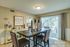 Inviting dining area with elegant decor, showcasing a wooden table with tall candle holders, cozy chairs, and a 'Stay awhile' wall sign. Natural light floods through large sliding glass doors leading to a serene outdoor view.