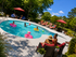 Inviting community pool area surrounded by lush greenery, featuring people enjoying a sunny day with loungers, umbrellas, and vibrant pool floats.