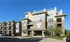 Modern apartment building with large windows and balconies, surrounded by lush greenery and ample parking space under a bright blue sky.