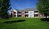 Charming multi-unit apartment building with classic brick facade, surrounded by lush green lawns and mature trees on a bright sunny day.