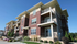 Modern multi-story apartment building with elegant balconies and charming brick exterior, surrounded by well-maintained greenery under a clear blue sky.