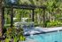 Luxurious outdoor patio area with modern pergola, dining set, and shimmering pool surrounded by lush palm trees.