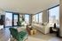 Bright and modern living room with expansive city views, featuring elegant green armchairs, a plush cream sofa, and stylish decor.