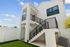 Modern two-story white home with stylish black railing staircase and lush green lawn, under a bright blue sky.