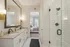 Elegant bathroom with a granite double sink vanity, stylish lighting, and a glass-enclosed shower; view into a cozy, sunlit bedroom with plush bedding.