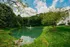 Serene apartment setting with lush green lawns, tranquil pond with fountain, and vibrant trees under a bright blue sky.