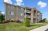Charming apartment building with beautiful landscaping, featuring a brick and siding exterior with balconies and lush green lawns under a bright blue sky.