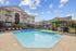Elegant apartment complex with a sparkling outdoor pool, surrounded by lush greenery and comfortable lounge chairs, under a bright blue sky.