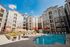 Modern apartment complex with a spacious courtyard, featuring a sparkling outdoor swimming pool, comfortable lounge chairs, and vibrant umbrellas under a sunny blue sky.