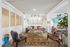 Elegant living room with stylish decor, featuring a cozy gray sofa adorned with patterned pillows, vibrant artwork, and a chic coffee table on a classic patterned rug. The space is brightened by natural light and includes modern lighting fixtures, diverse seating options, and lush greenery.