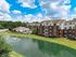 Charming apartment complex with a serene lakeside view, sparkling pool, and vibrant red brick architecture under a bright blue sky.