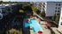 Modern apartment complex with a large swimming pool, surrounded by stylish outdoor seating and vibrant red umbrellas, set against a backdrop of lush green trees and contemporary buildings.