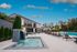 Luxurious outdoor pool area with modern clubhouse, cascading waterfall feature, and inviting lounge chairs under vibrant umbrellas set against a clear blue sky.